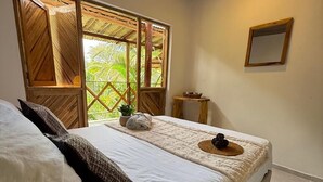 A bedroom with a bed, pillows, a small table, a mirror, and a window with a view of greenery. - Zelandia Eco Finca (Santa Marta)