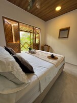 Un dormitorio con cama, almohadas, una mesita, un espejo y una ventana con vistas a la vegetación.