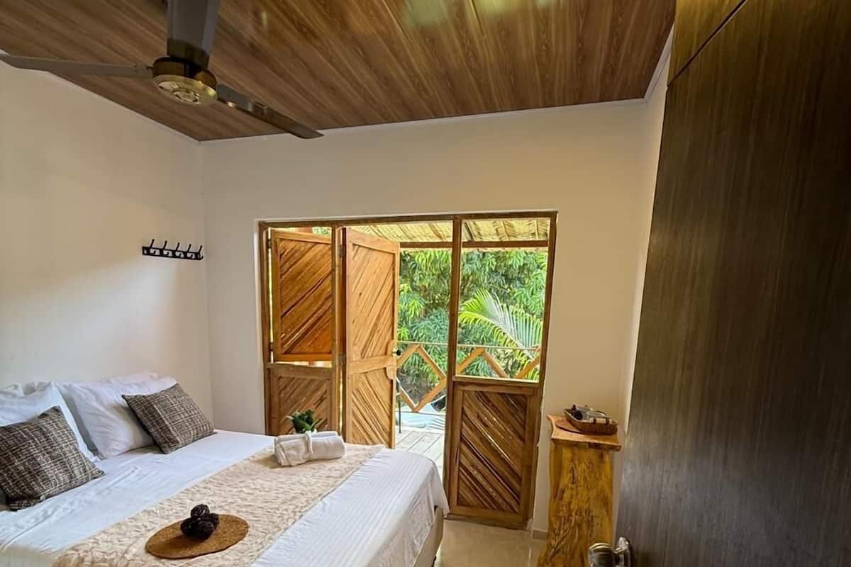 Une chambre à coucher avec un lit, un ventilateur de plafond, une fenêtre donnant sur un paysage verdoyant et une armoire en bois.
