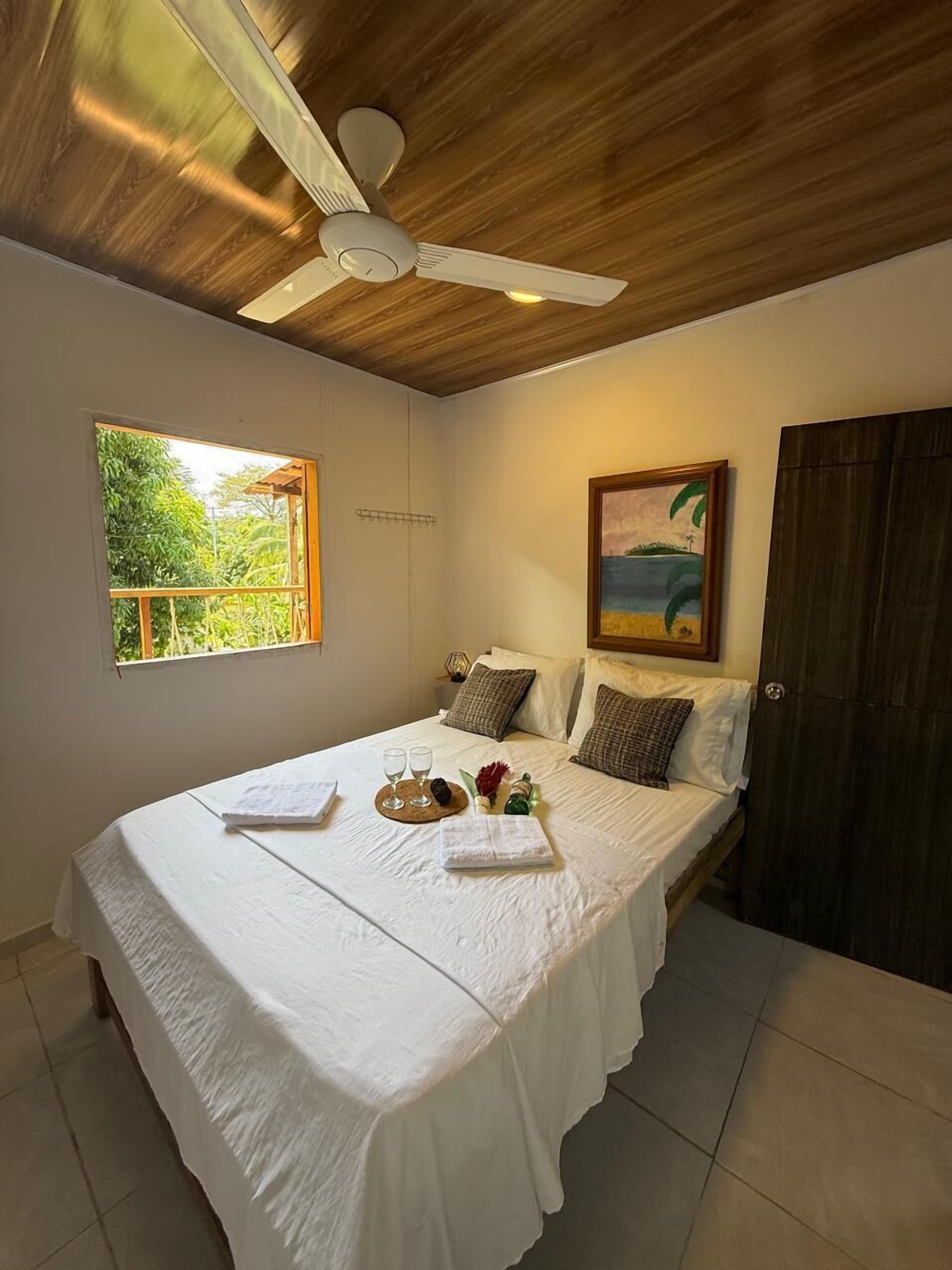 Un dormitorio con cama, ventilador de techo, ventana con vistas, un cuadro y una puerta.
