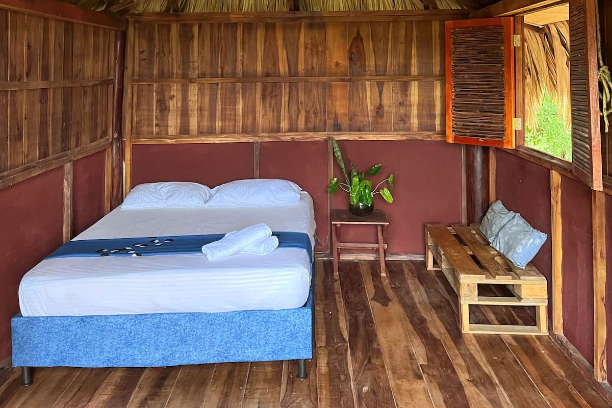 Une pièce comprenant un lit, un banc, un ventilateur de plafond et une fenêtre donnant sur un paysage verdoyant.