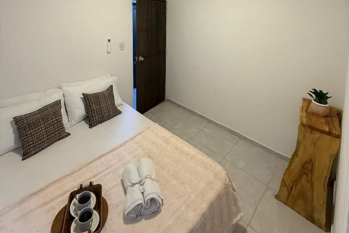 Une chambre avec un plafond en bois, un lit recouvert d’une couverture beige, une petite table de chevet en bois ornée d’une plante, et une porte donnant sur une autre pièce.