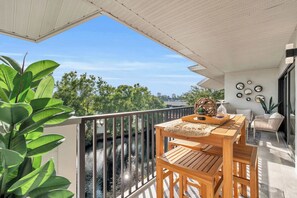 A balcony with a wooden table and chairs, a potted plant, and a view of a river and cityscape.