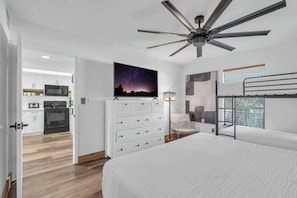 A bedroom with a bed, a ceiling fan, a dresser, a nightstand, a lamp, a bunk bed, and a view of the kitchen. - I-452 - Remodeled, Luxury Condo with Lake Views (Naples)