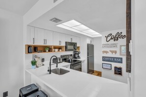 A modern kitchen with white cabinetry, a built-in microwave, a coffee station, and various kitchen appliances. - I-452 - Remodeled, Luxury Condo with Lake Views (Naples)