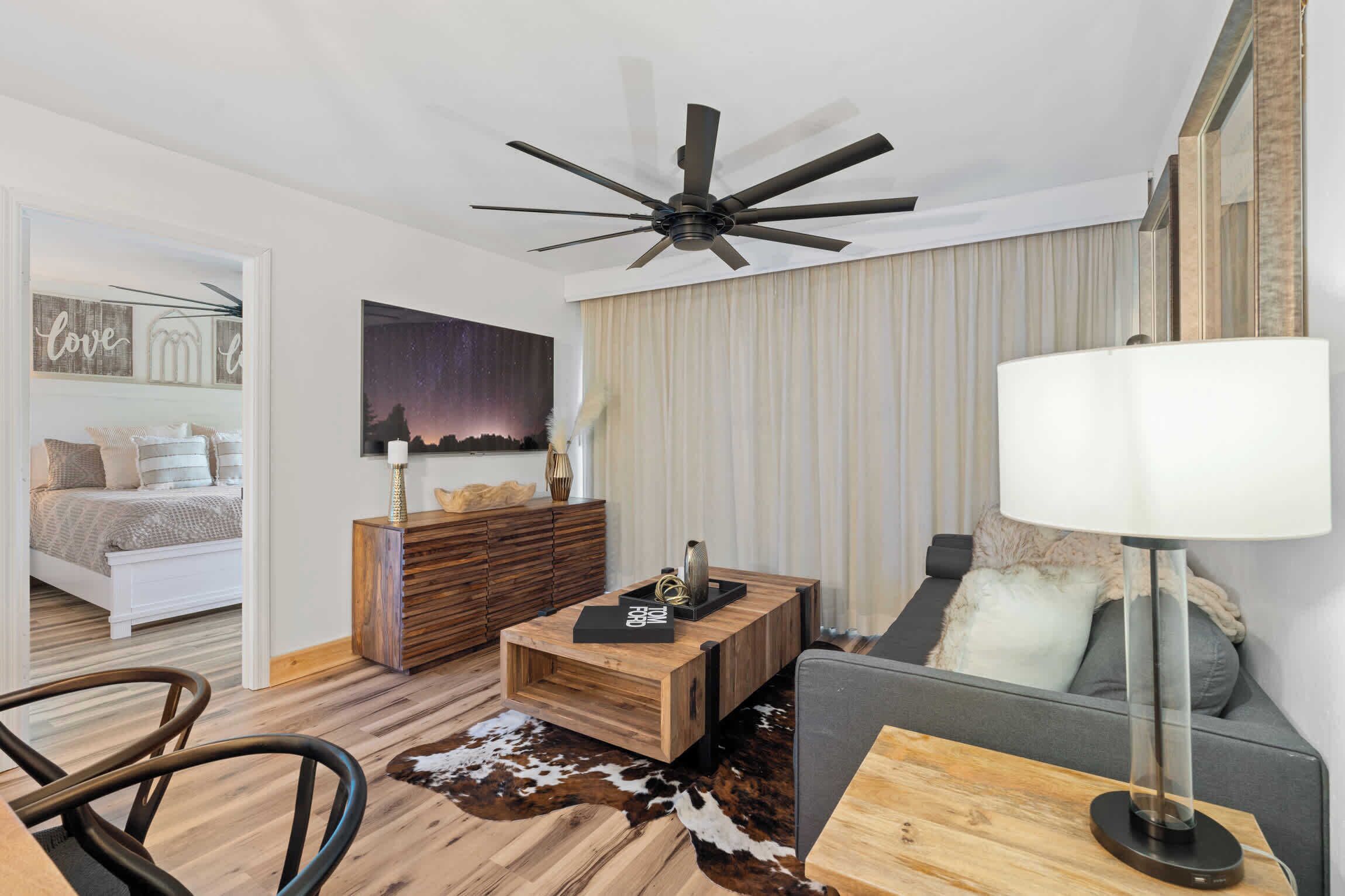 Une chambre d’hôtel moderne avec un lit, une table basse en bois, un canapé, un ventilateur de plafond et une grande fenêtre avec des rideaux.