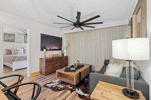 Une chambre d’hôtel moderne avec un lit, une table basse en bois, un canapé, un ventilateur de plafond et une grande fenêtre avec des rideaux.