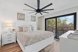 A bedroom with a large bed, a ceiling fan, a nightstand with a lamp, and a view of the outdoors through a sliding glass door.