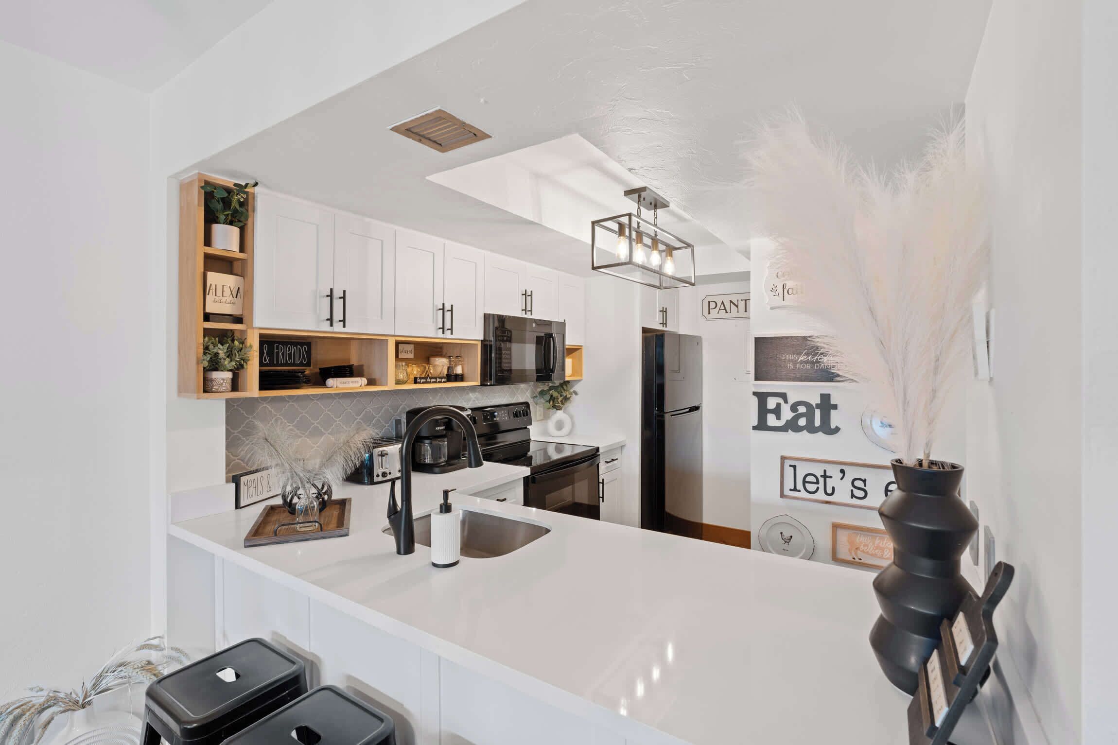 Une cuisine moderne avec des meubles blancs, un réfrigérateur noir et un plan de travail blanc. On peut voir un vase noir orné de plumes blanches posé sur le plan de travail.