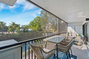 A balcony with wicker furniture, a glass table, and a view of a water body and trees.