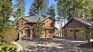 A two-story wooden cabin with a stone foundation, a garage, and a driveway surrounded by trees. - Walk to the Winery & Chip onto the Par 3 Course! (Cle Elum)