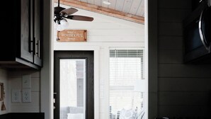 A modern kitchen with dark cabinetry, a stove, and a ceiling fan. A cozy seating area with a gray sofa and patterned cushions is visible through a doorway. - Be Still Tiny House on Monteagle Mountain  (Tracy City)