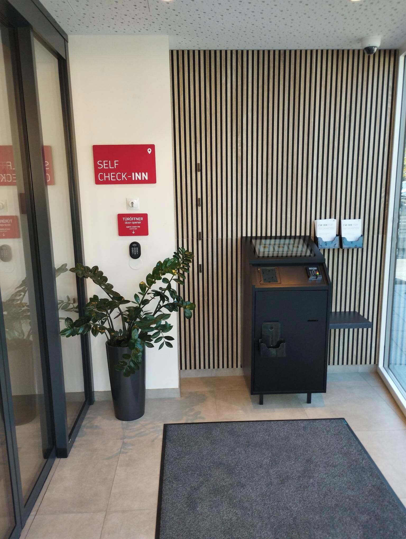 Ein Empfangsbereich mit Schilder für Selbst-Check-in, einem Türöffner, einem Blumentopf, einer Überwachungskamera und einem kleinen Schreibtisch mit einem Computer.