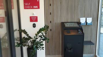 Ein Empfangsbereich mit Schilder für Selbst-Check-in, einem Türöffner, einem Blumentopf, einer Überwachungskamera und einem kleinen Schreibtisch mit einem Computer.