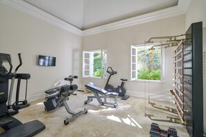Une salle de sport à domicile équipée de matériel d’exercice, d’un téléviseur et de fenêtres donnant sur un paysage verdoyant.