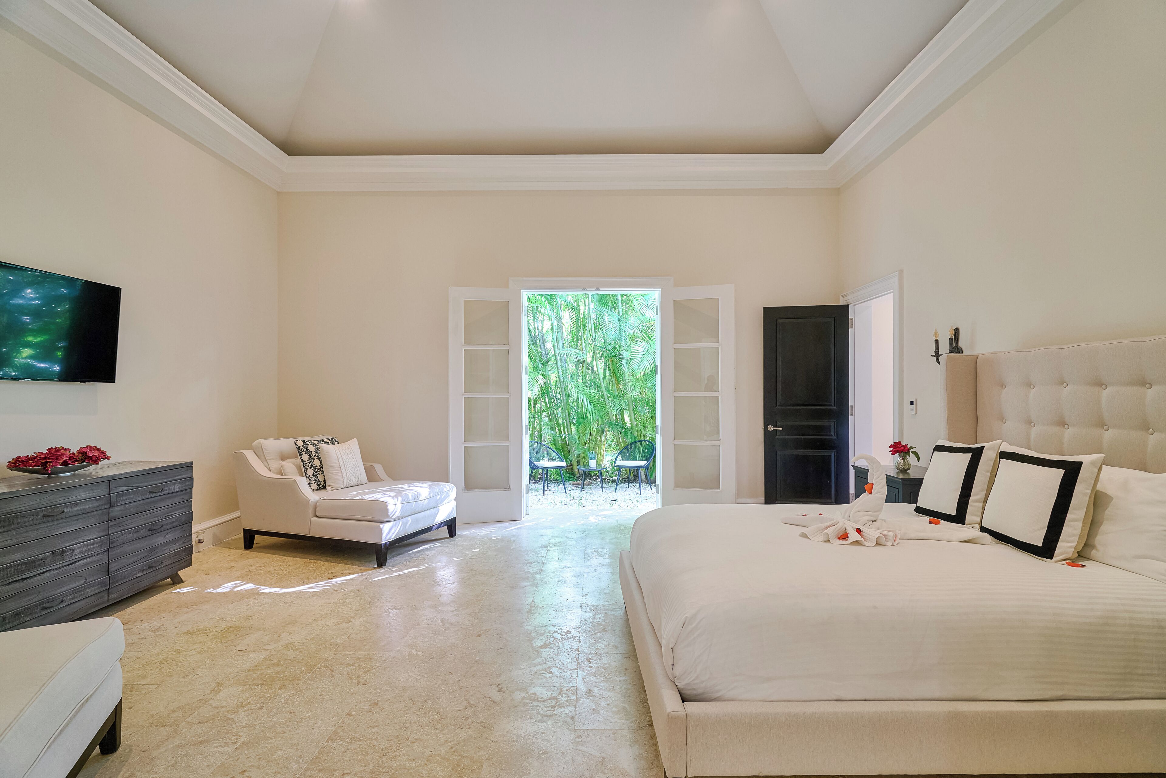 Quarto com cama, cadeira, cômoda, televisão e porta que dá para um jardim.