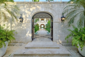 A stone archway leading to a courtyard with a pool, surrounded by palm trees and a building with balconies.