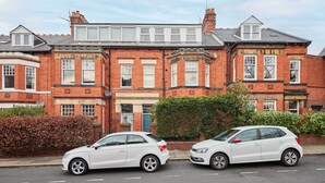 A row of red brick houses with white windows and doors, parked cars on the street, and a clear sky. - Host & Stay | The Rooftop Gallery (Jesmond)