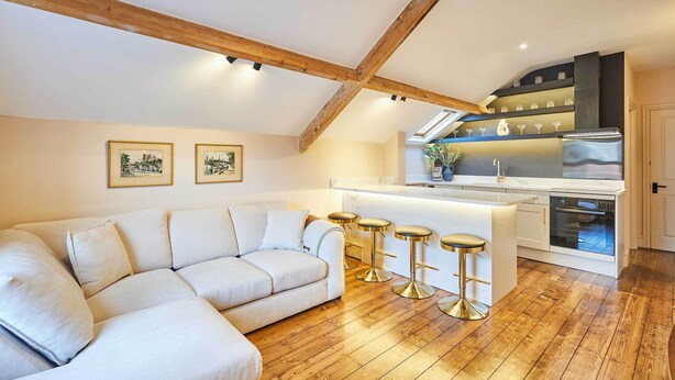 A modern loft with a wooden ceiling, a white sofa, a kitchen with a bar area, and two framed pictures on the wall. - Host & Stay | The Rooftop Gallery (Jesmond)