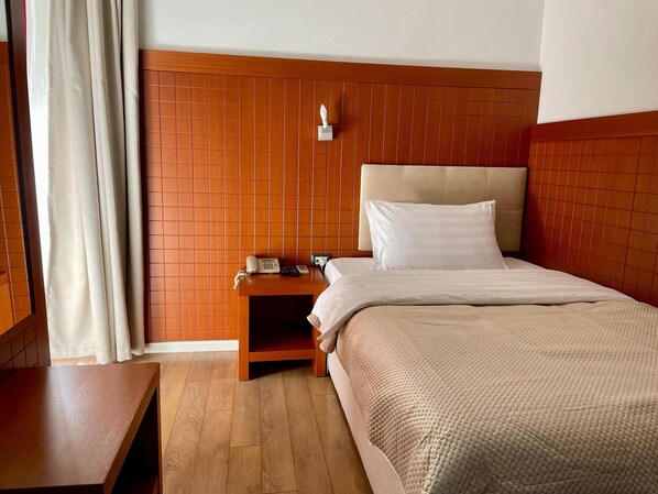 A hotel room with a wooden headboard, a wooden nightstand with a telephone and a lamp, a bed with white bedding, and a wooden floor. - City 5 Hotel (Tirana)