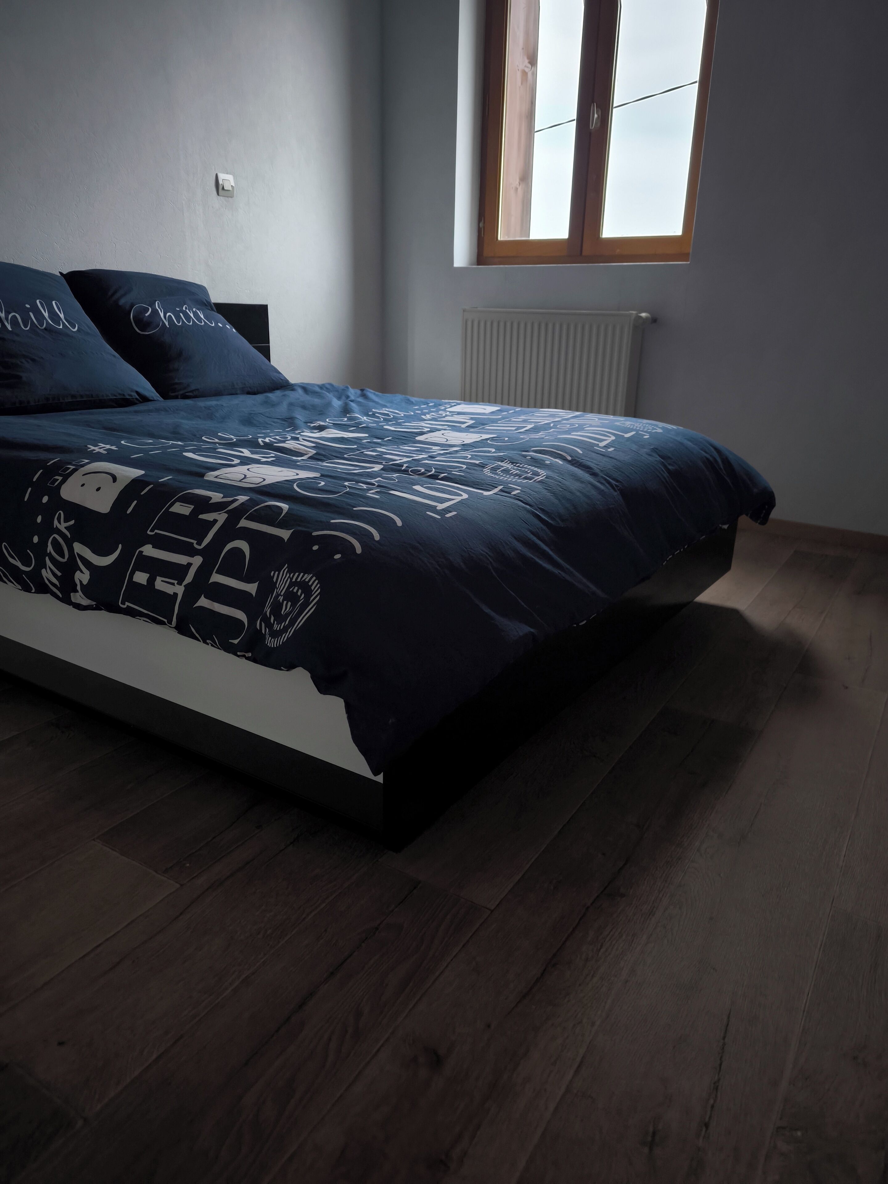 Une chambre à coucher avec un lit, un parquet en bois foncé, une fenêtre et un radiateur.