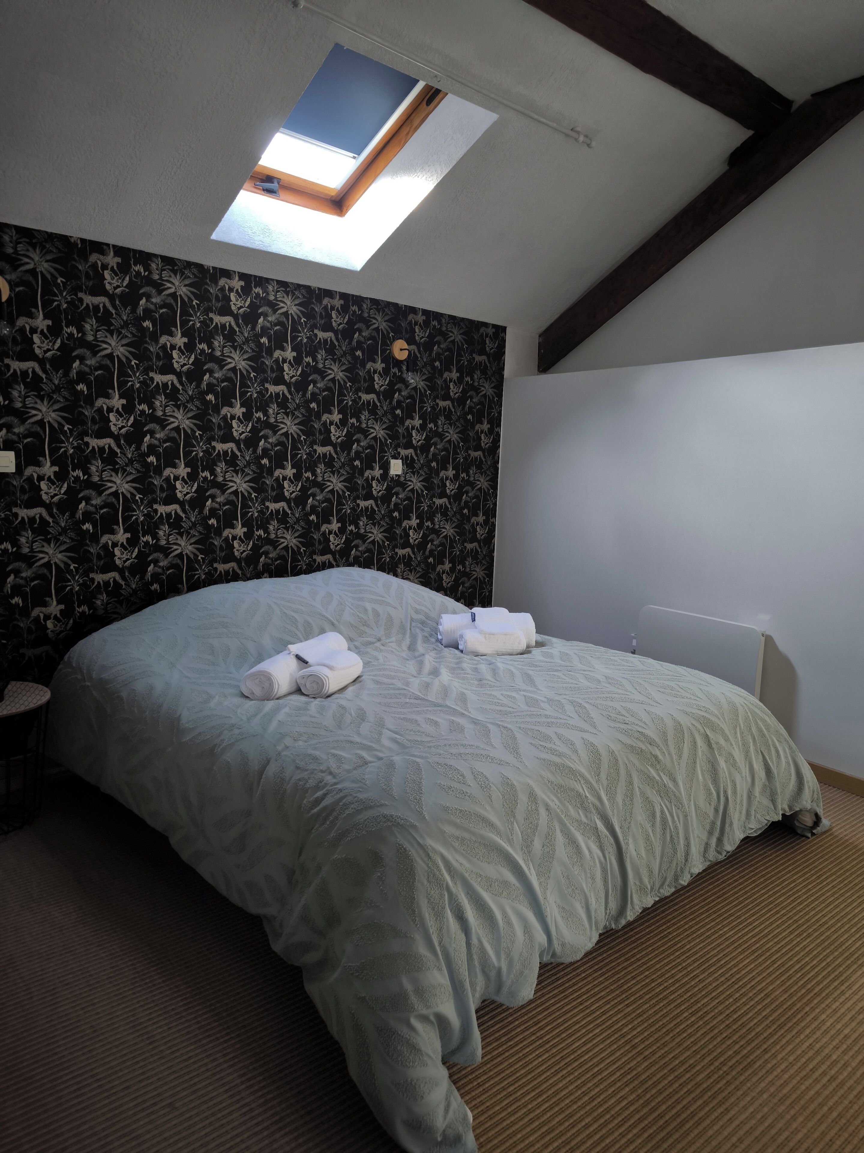 A bedroom with a sloped ceiling, a skylight, a patterned wallpaper, a bed with a white bedspread, and rolled towels on the bed.