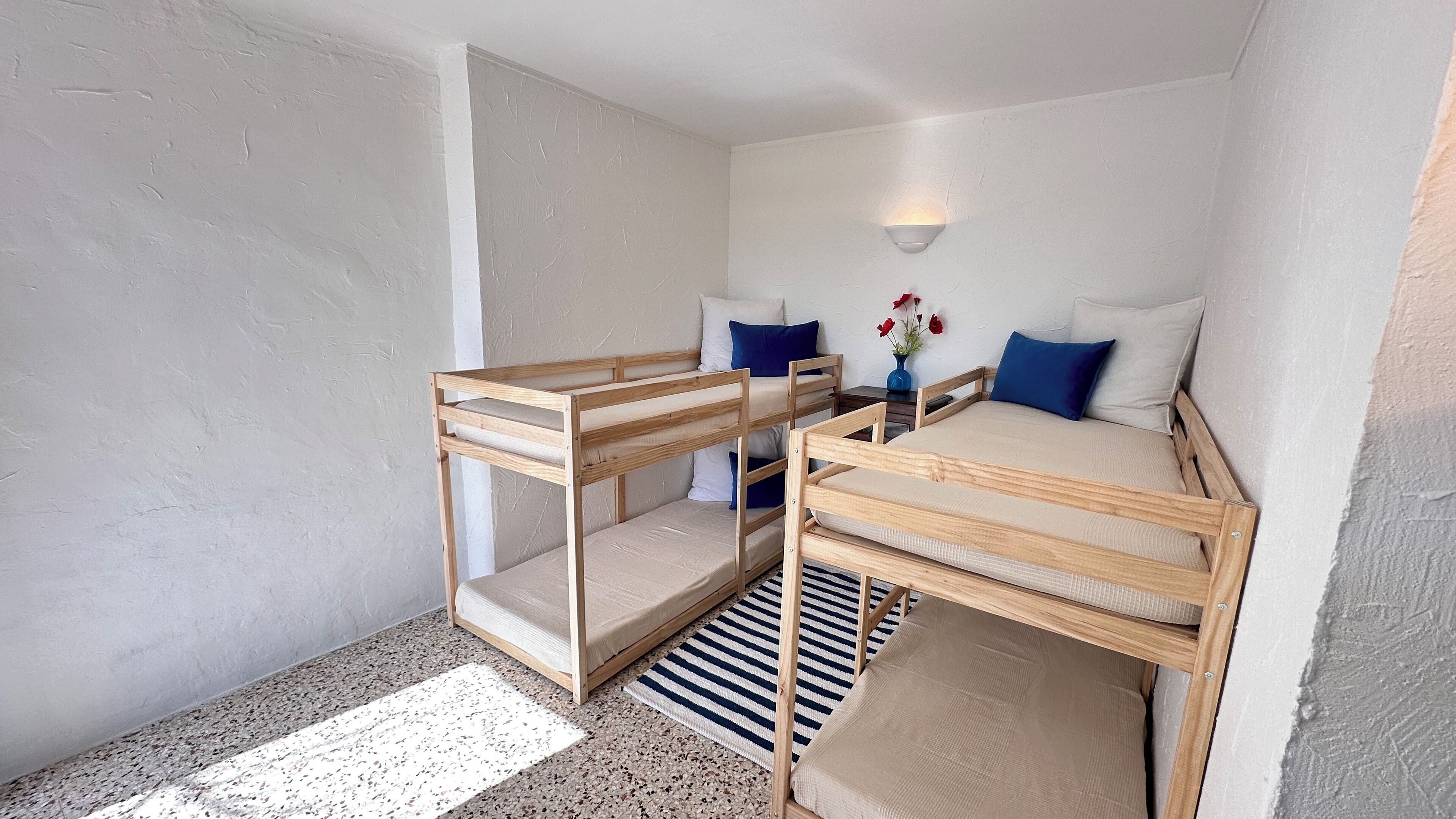 Une petite chambre avec des lits superposés, une petite table de chevet ornée d’un vase de fleurs et un luminaire fixé au mur.