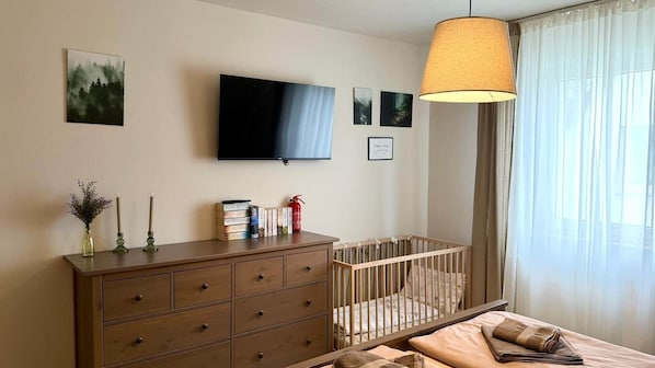 Un dormitorio con una cuna, un televisor montado en la pared, un aparador y una ventana con cortinas de gasa.
