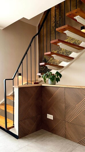 Una escalera con peldaños de madera y barandillas negras, una planta en maceta en la plataforma, y un papel tapiz decorado.