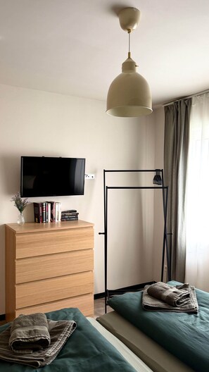 A room with a TV, wooden dresser, books, a standing fan, and a bed with towels. - Breathing space-Spreewald (Vetschau/Spreewald)