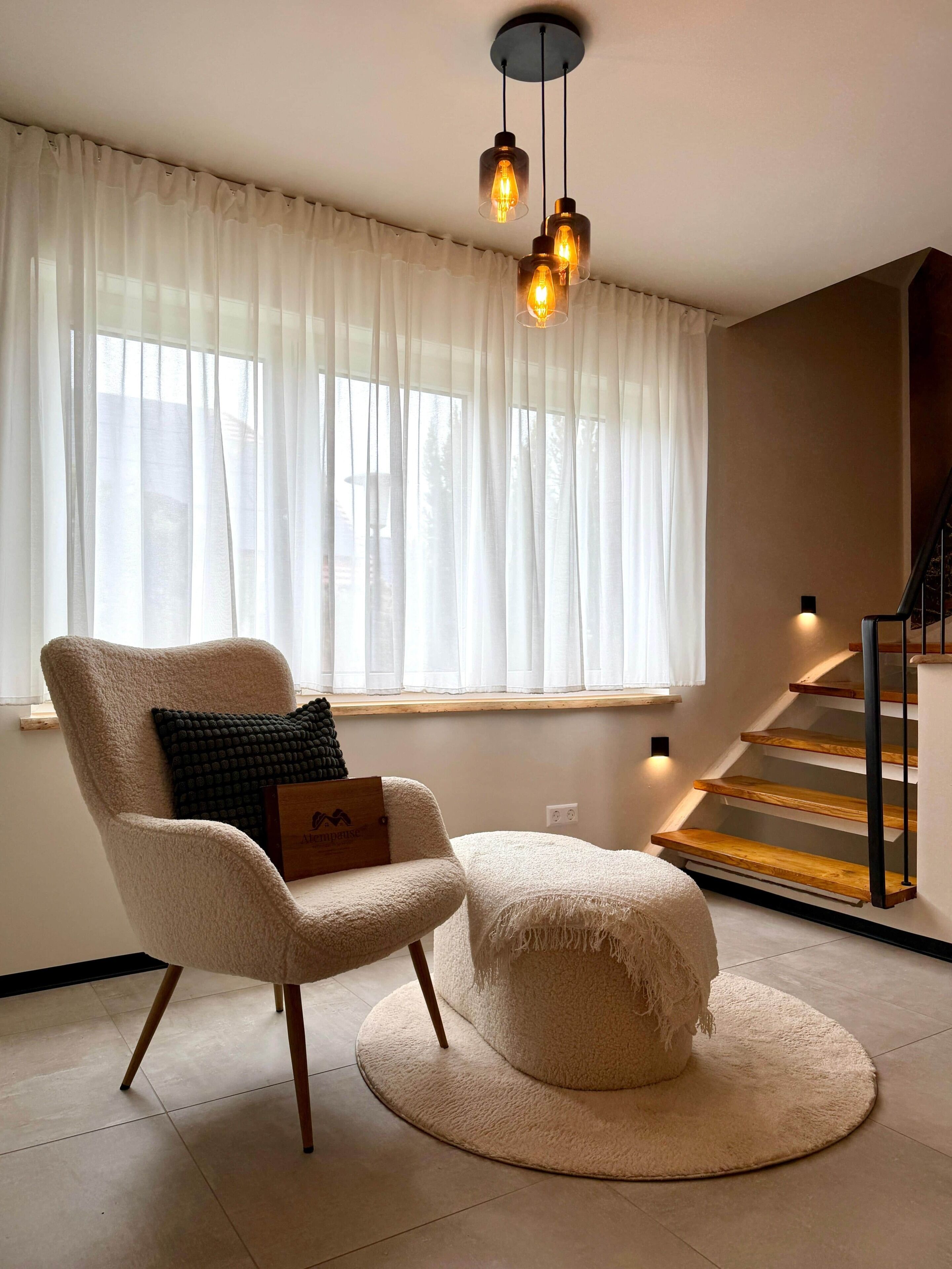 A modern living room with a large window, a chair, a round ottoman, and a staircase.