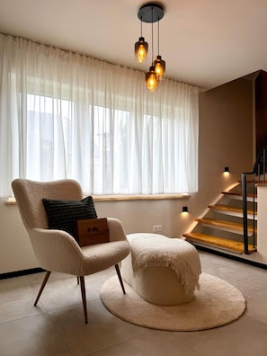 A modern living room with a large window, a chair, a round ottoman, and a staircase. - Breathing space-Spreewald (Vetschau/Spreewald)