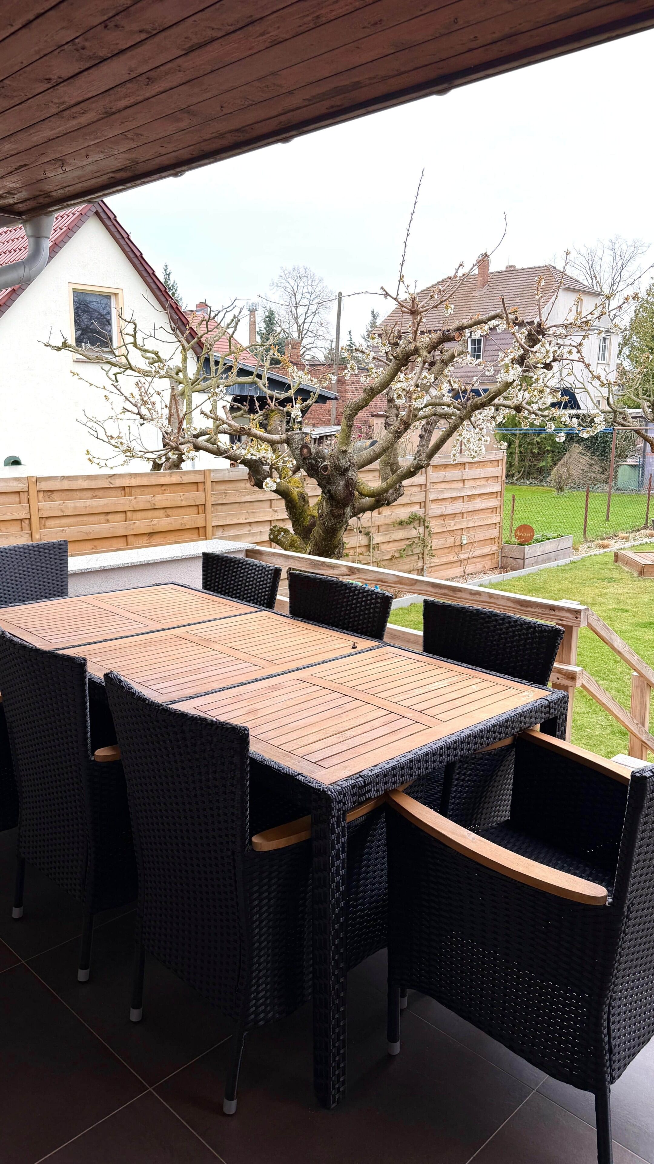 Un patio al aire libre con una mesa de madera y sillas de mimbre, un árbol en flor y vistas de casas al fondo.