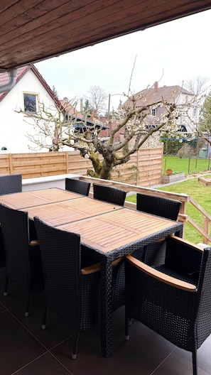 Un patio al aire libre con una mesa de madera y sillas de mimbre, un árbol en flor y vistas de casas al fondo.