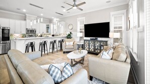 A modern kitchen with white cabinets, a large island, and bar stools. There is a living area with a beige sofa, a coffee table, and a TV. - 30A House with Pool and Bunk Rooms (Seacrest Beach)