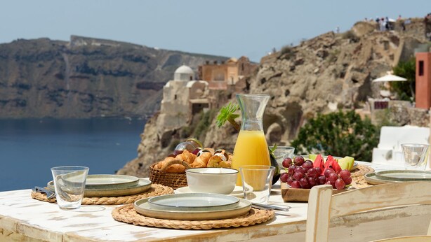 A table set for breakfast with plates, glasses, and a fruit basket, overlooking a coastal landscape with buildings and cliffs. - Ilivatos - Elegant Villa on the Caldera's Edge (Oia)