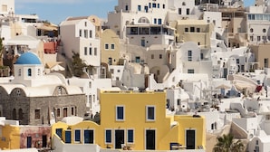A densely packed hillside of white buildings with a prominent yellow structure in the center, surrounded by steep staircases and palm trees. - Ilivatos - Elegant Villa on the Caldera's Edge (Oia)