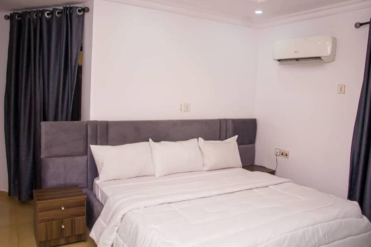 A bedroom with a large bed, a nightstand, a wall-mounted air conditioner, and dark curtains.