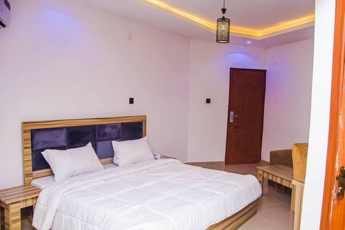 A bedroom with a large bed, wooden headboard, bedside tables, a hanging light fixture, and a door in the background.