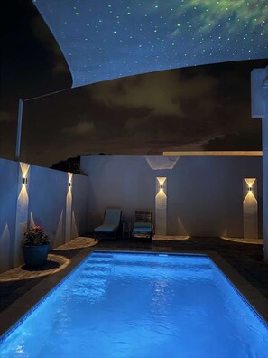 A swimming pool with a starry sky above, surrounded by walls with built-in lighting, a lounge chair, and a potted plant.