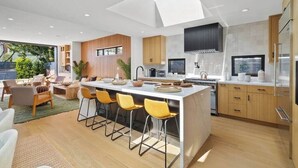 A modern kitchen with a central island, bar stools, and a large window overlooking a garden. - Venice Open Designer House w Pool (Los Angeles)