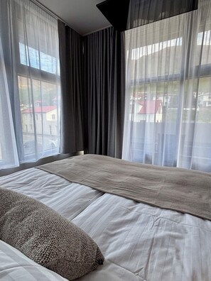 A modern hotel room with a large bed, sheer curtains, and a view of buildings outside.