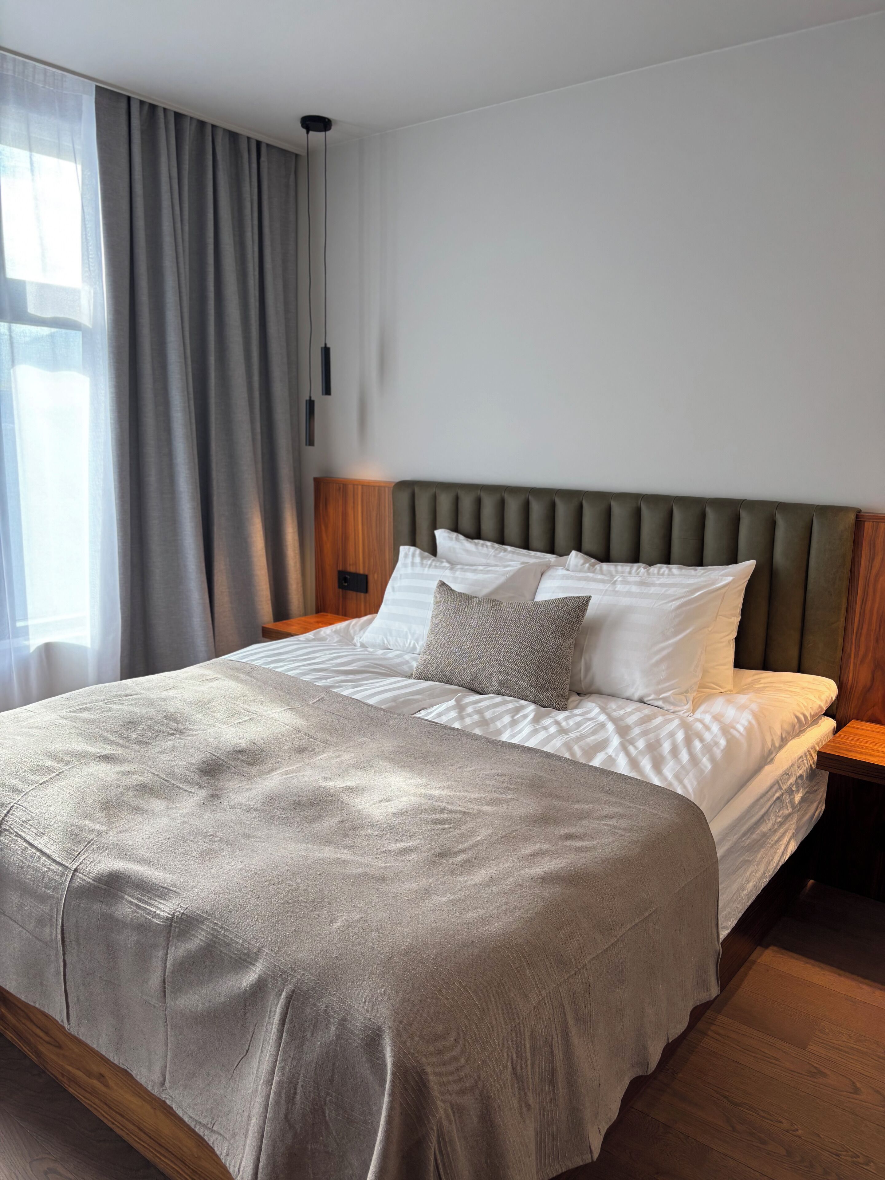 A modern bedroom with a large bed, grey curtains, and a wooden nightstand.