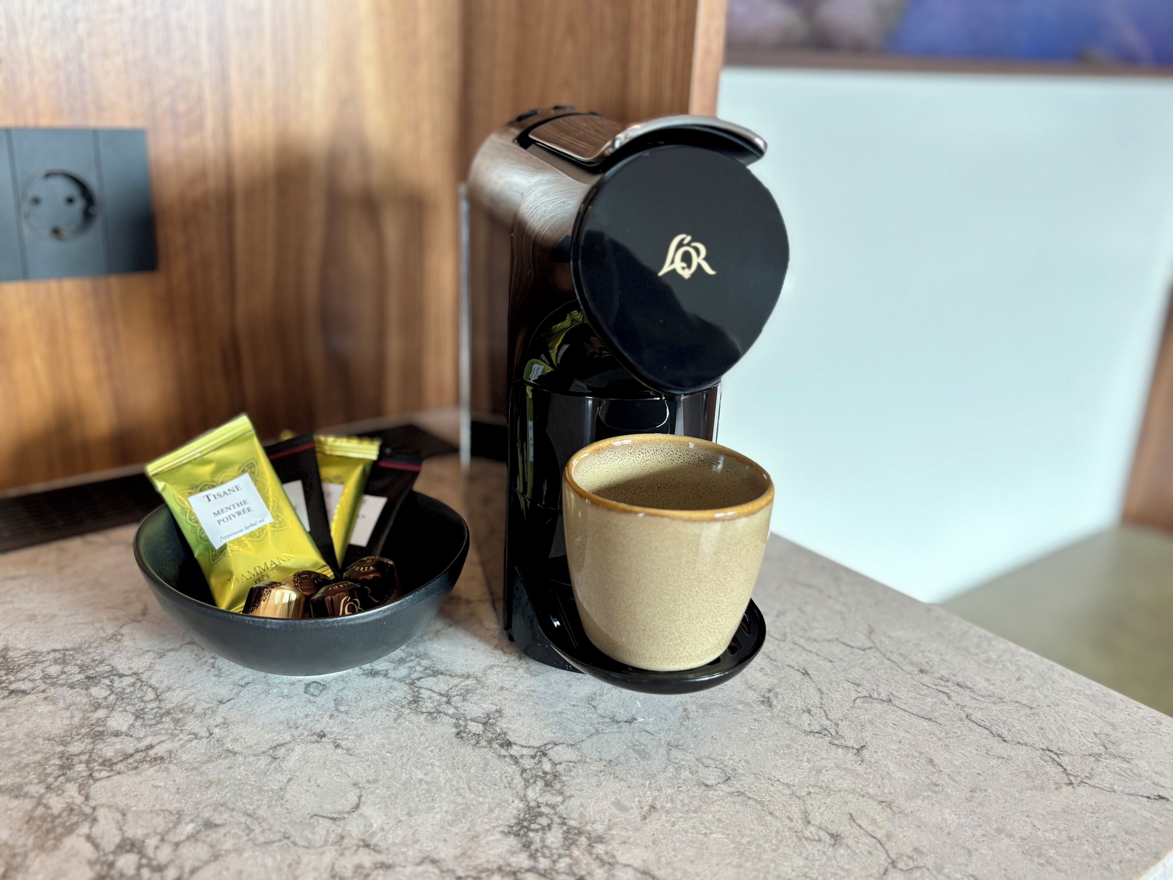 A coffee maker with a cup and a bowl containing coffee pods and a spoon on a marble countertop.