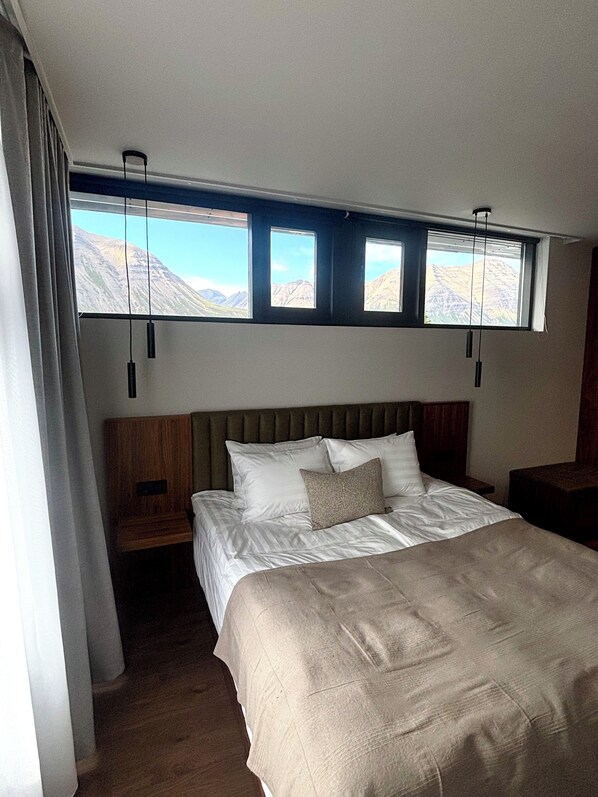 A bedroom with a large bed, a wooden headboard, and a view of mountains through three windows.