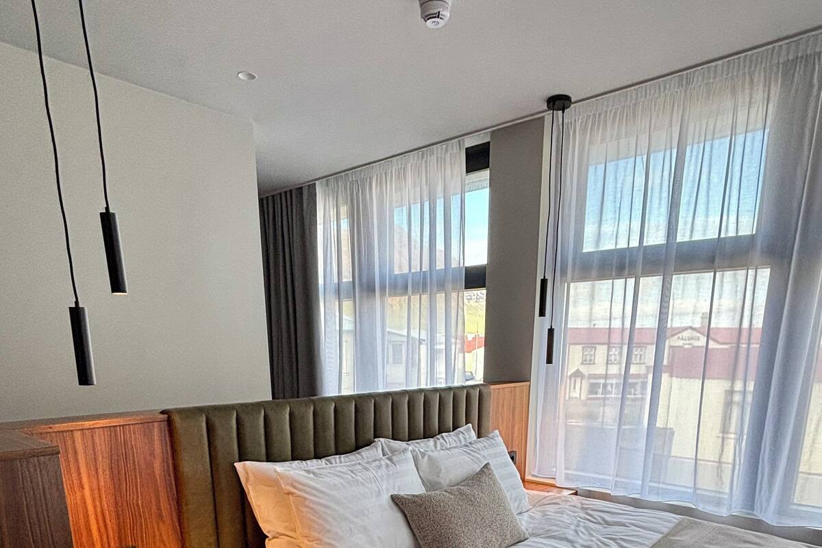 Una habitación de hotel moderna con una cama grande, cabecero de madera y vistas al exterior a través de cortinas translúcidas.