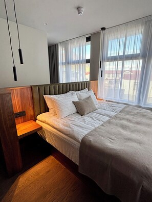 A modern hotel room with a large bed, wooden headboard, and a view of the outside through sheer curtains.
