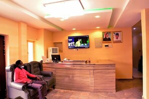 A reception area with a wooden reception desk, a television mounted on the wall, and two leather chairs. - Mimantex Apartment (Abuja)