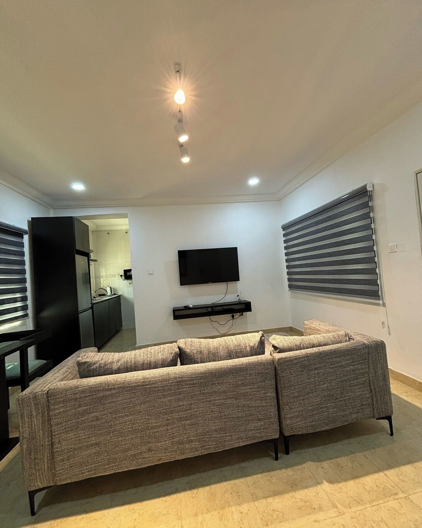 A living room with a sofa, a TV mounted on the wall, and a kitchen area visible through an open door.