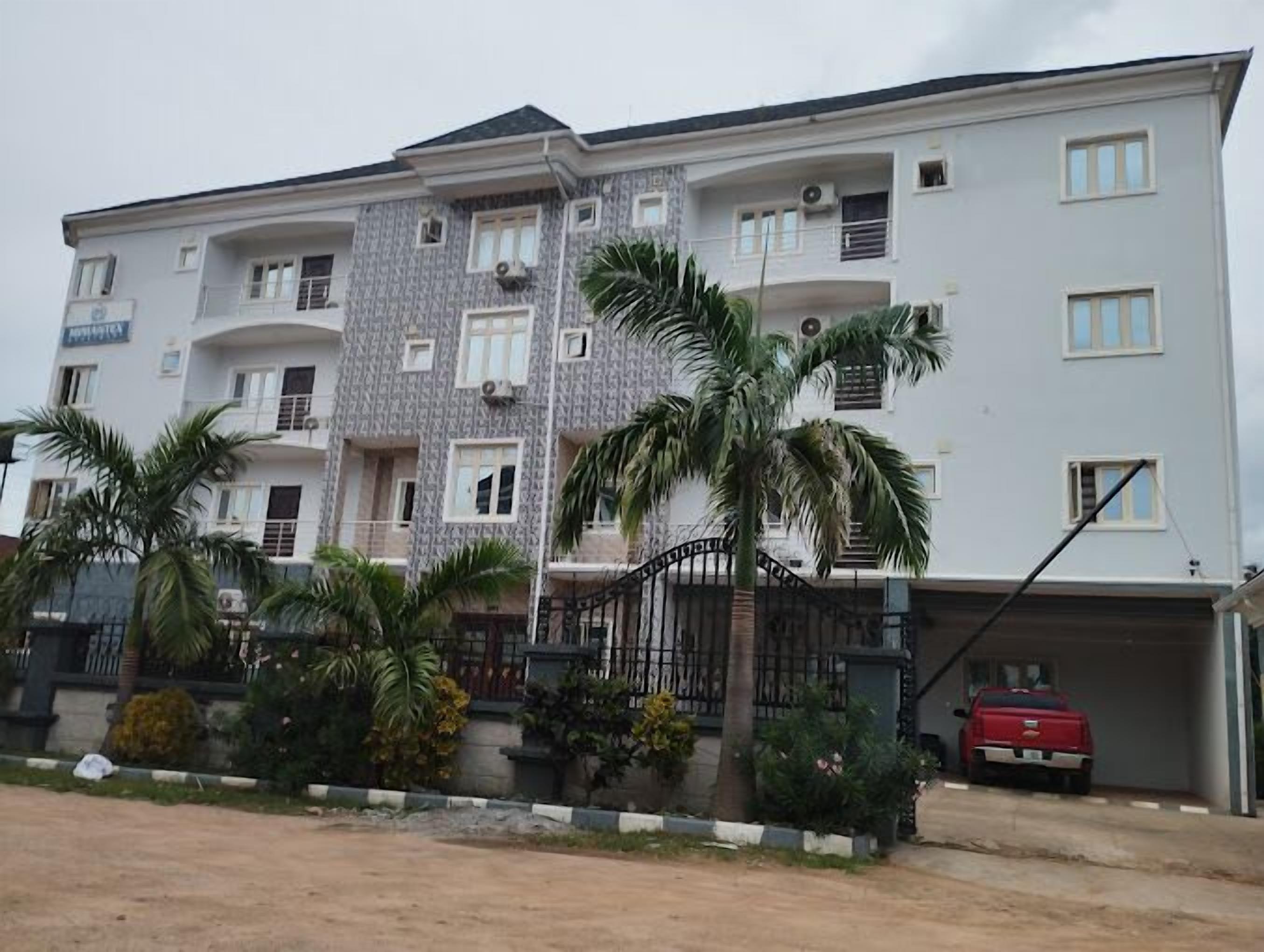 A multi-story residential building with balconies, a gated entrance, and a red vehicle parked in the driveway.